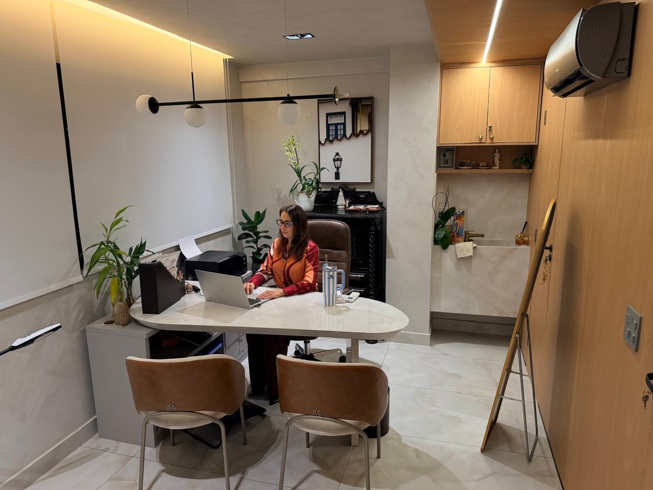 Dra. Avany à mesa em sua sala de atendimento, com poltronas e decoração em tons claros e madeira